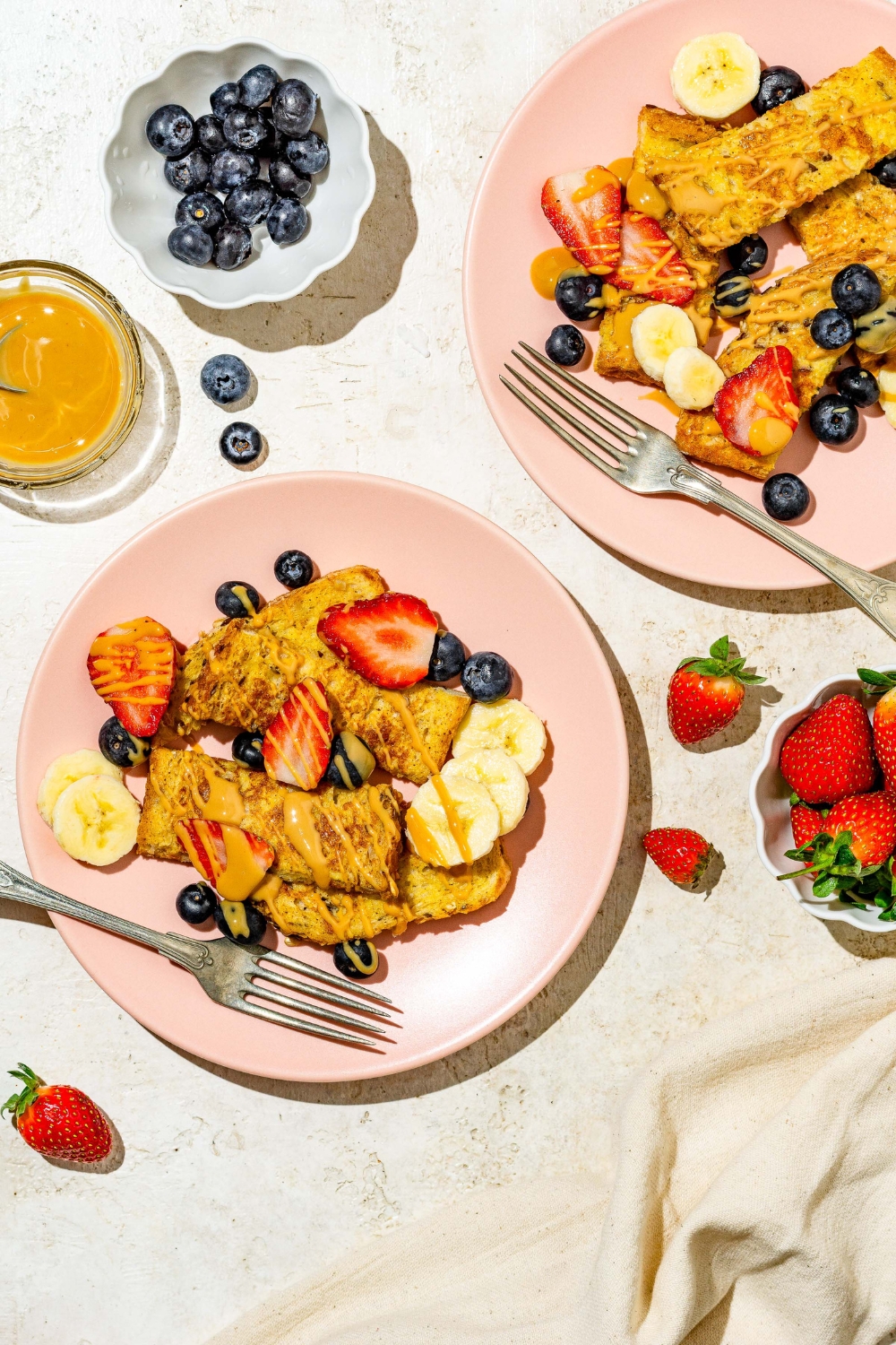 A pink plate with sliced baby french toast sticks toped with sliced banana, blueberries, and strawberries and drizzled with peanut butter. There is a fork on the plate. The plate is on a tan counter with an additional plate of french toast and small bowls of toppings.