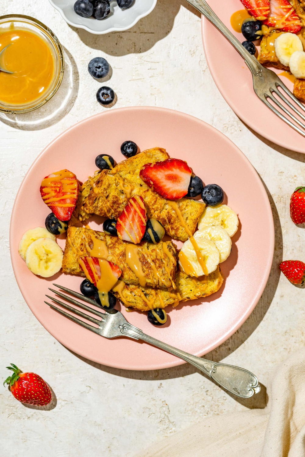 A pink plate with sliced baby french toast sticks toped with sliced banana, blueberries, and strawberries and drizzled with peanut butter. There is a fork on the plate. The plate is on a tan counter with an additional plate of french toast and small bowls of toppings.