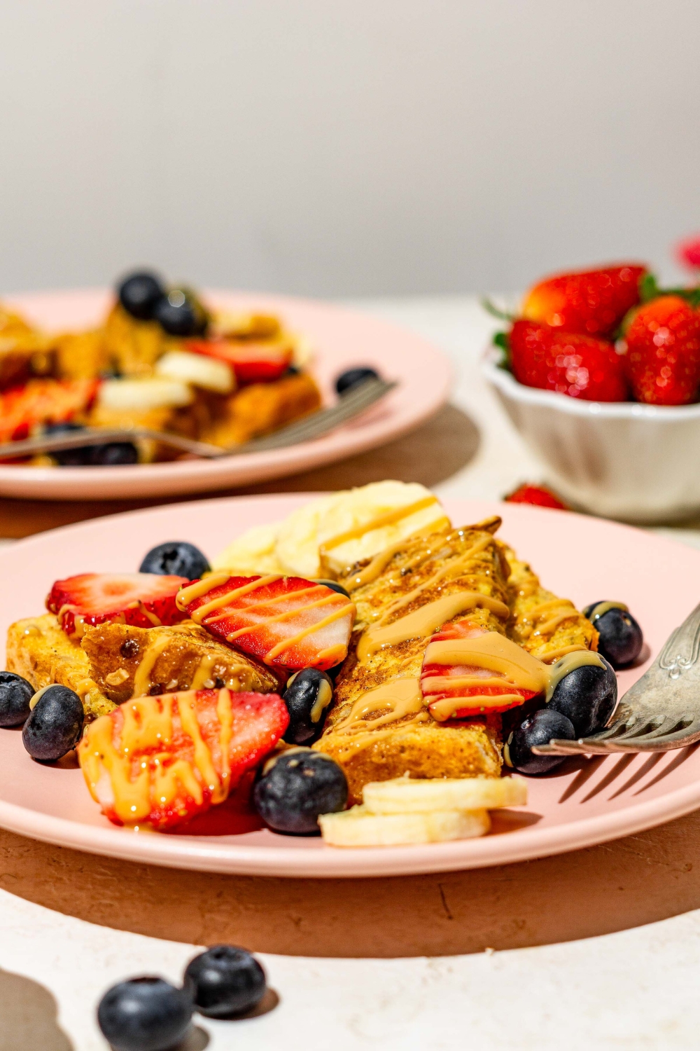 A pink plate with sliced baby french toast sticks toped with sliced banana, blueberries, and strawberries and drizzled with peanut butter. There is a fork on the plate. The plate is on a tan counter with an additional plate of french toast and small bowl of strawberries.