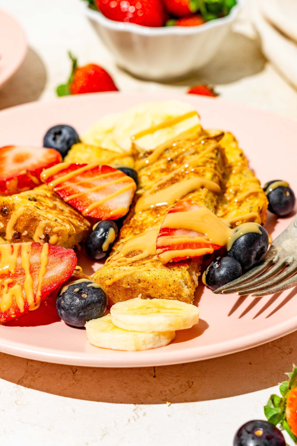 A pink plate with sliced baby french toast sticks toped with sliced banana, blueberries, and strawberries and drizzled with peanut butter. There is a fork on the plate. The plate is on a tan counter with an additional plate of french toast and small bowl of blueberries.