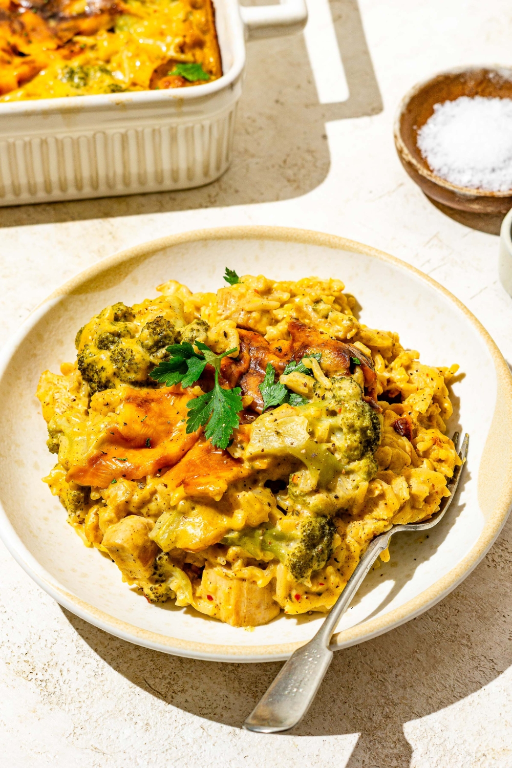 A bowl with chicken broccoli rice casserole garnished with fresh parsley. There is a fork in the bowl. The bowl is on a tan counter with a dish of casserole.