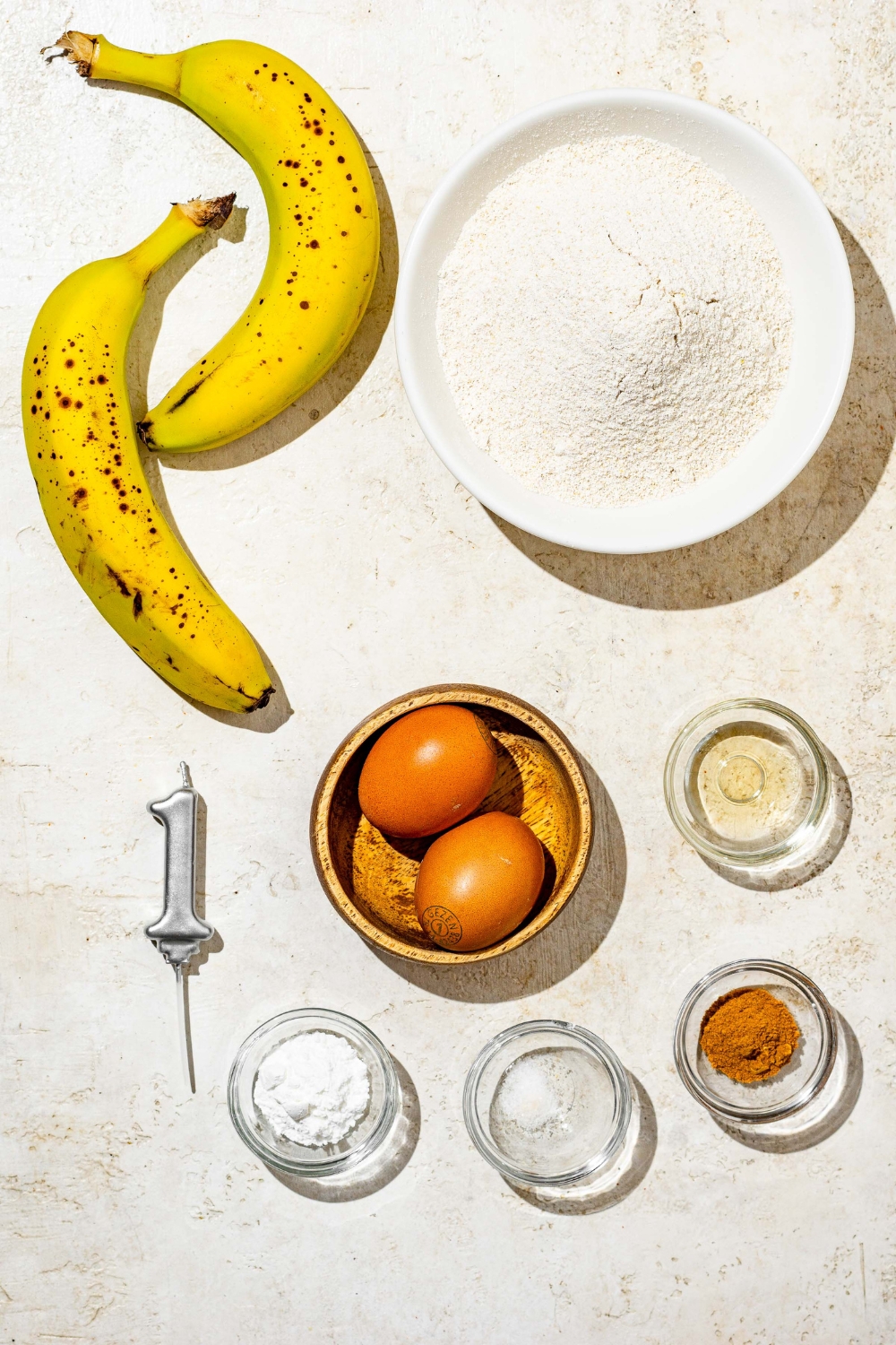 An overhead shot of several bowls in various sizes containing ingredients to make banana smash cake including bananas, almond flour, eggs, baking powder, cinnamon, and vanilla extract.