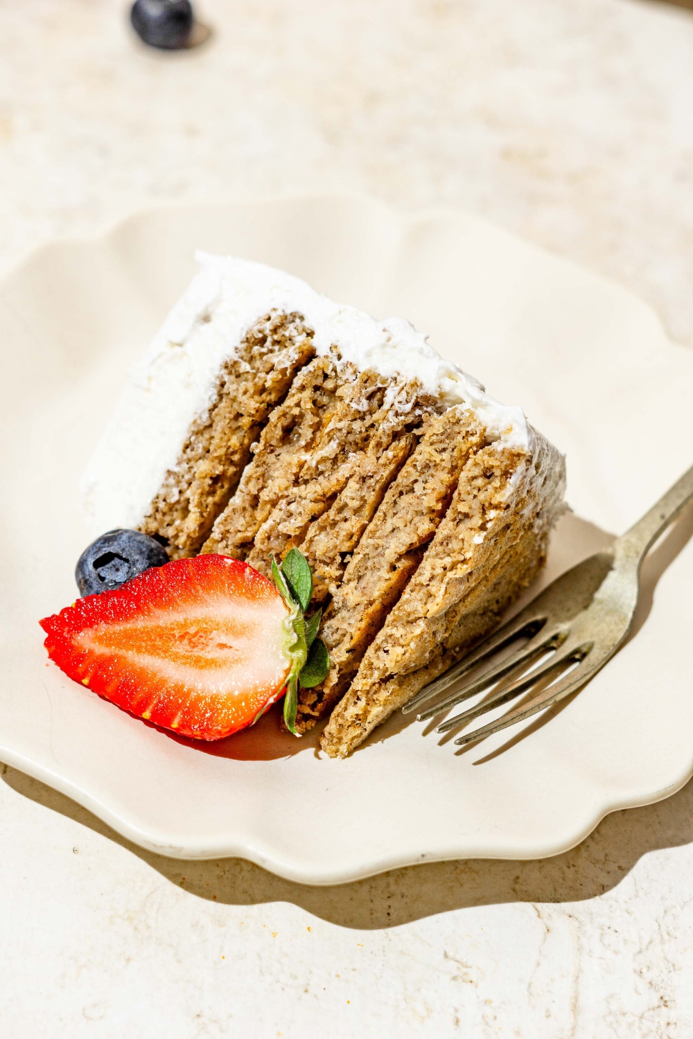 A slice of frosted banana smash cake on a white scalloped plate with a fork and sliced strawberry. The plate is on a tan counter.