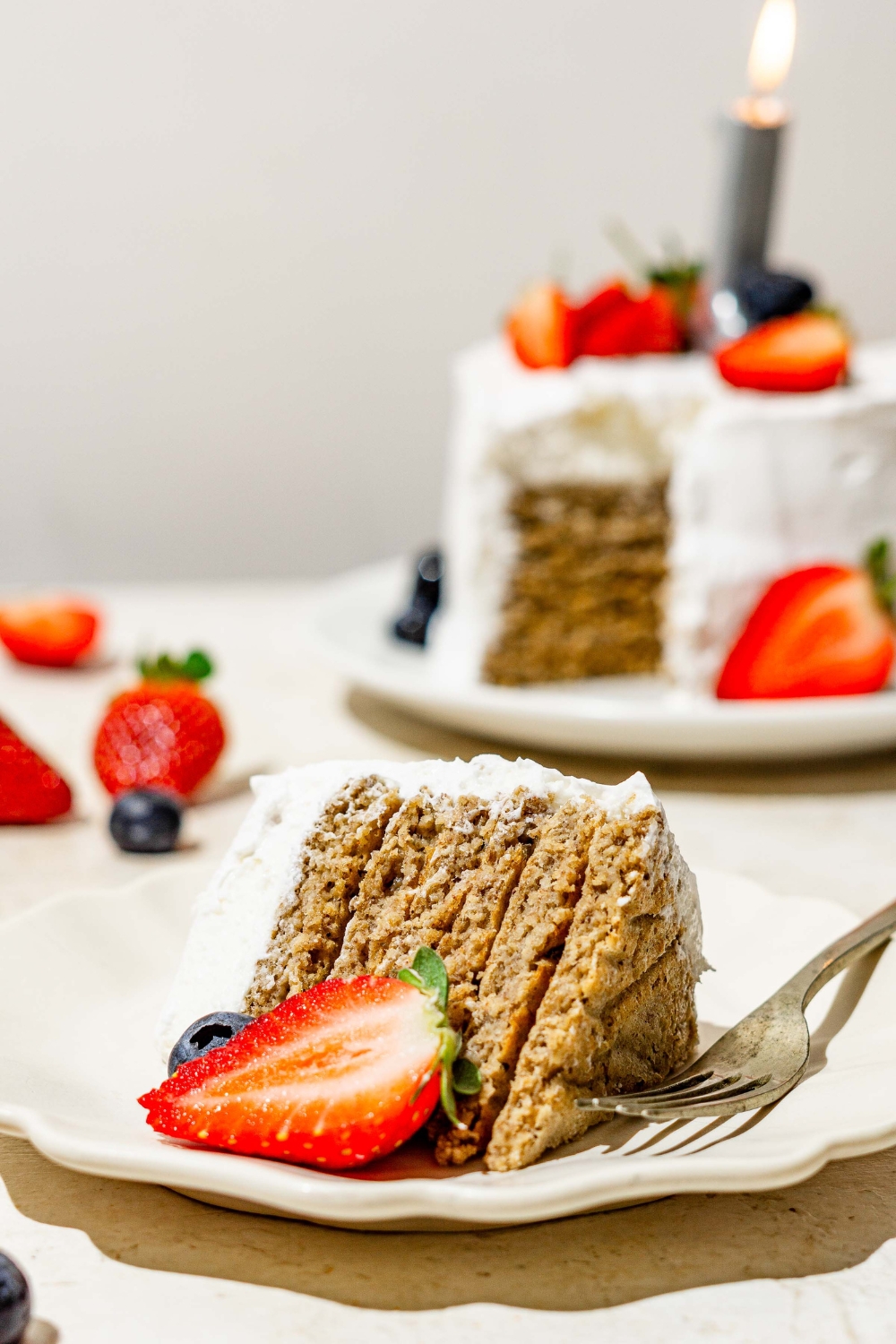 A slice of frosted banana smash cake on a white scalloped plate with a fork and sliced strawberry. The plate is on a tan counter with a whole banana smash cake.