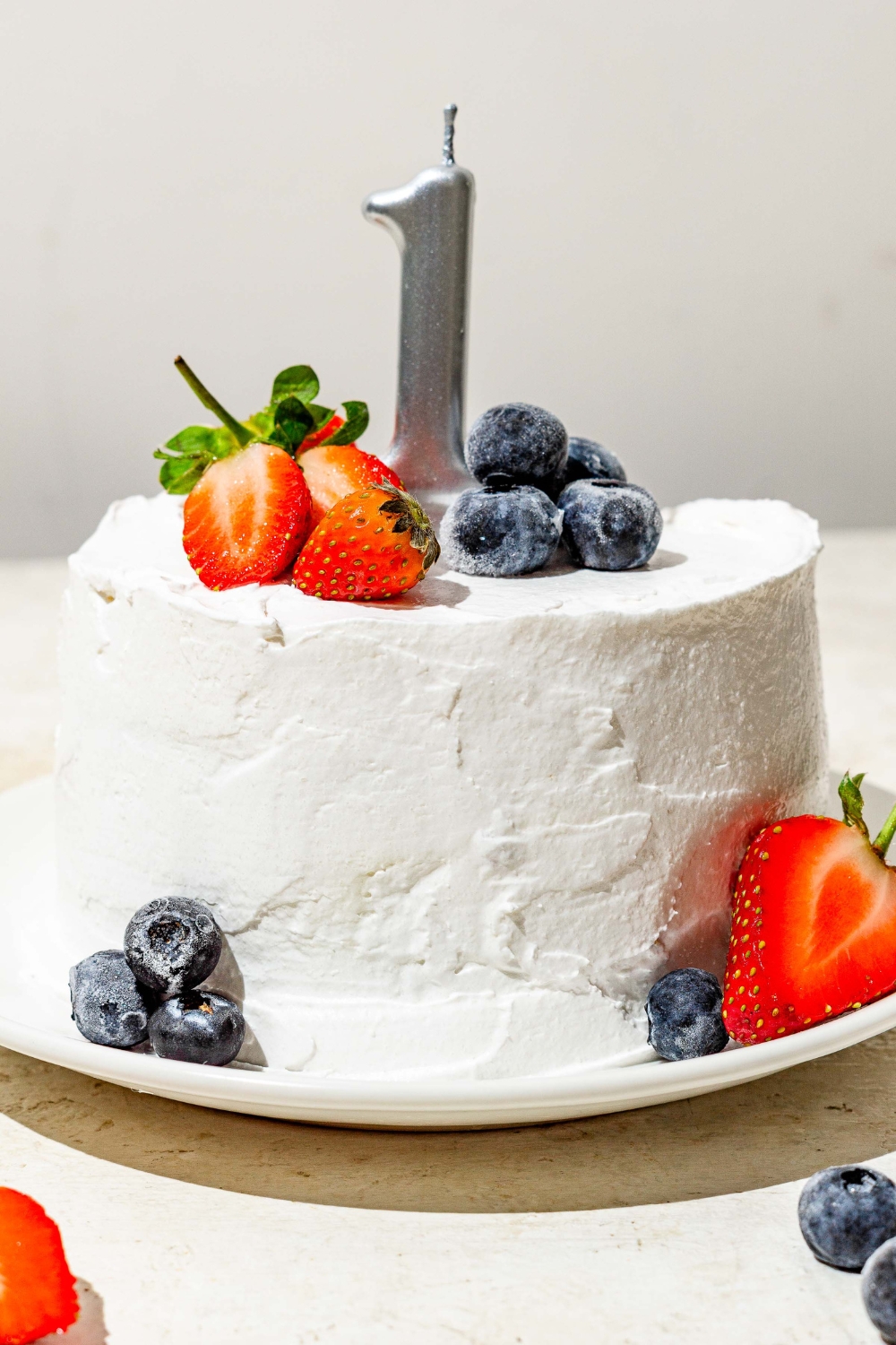 A frosted banana smash cake topped with strawberries, blueberries, and a birthday candle of the number 1. The cake is on a white plate on a tan counter with a knife.