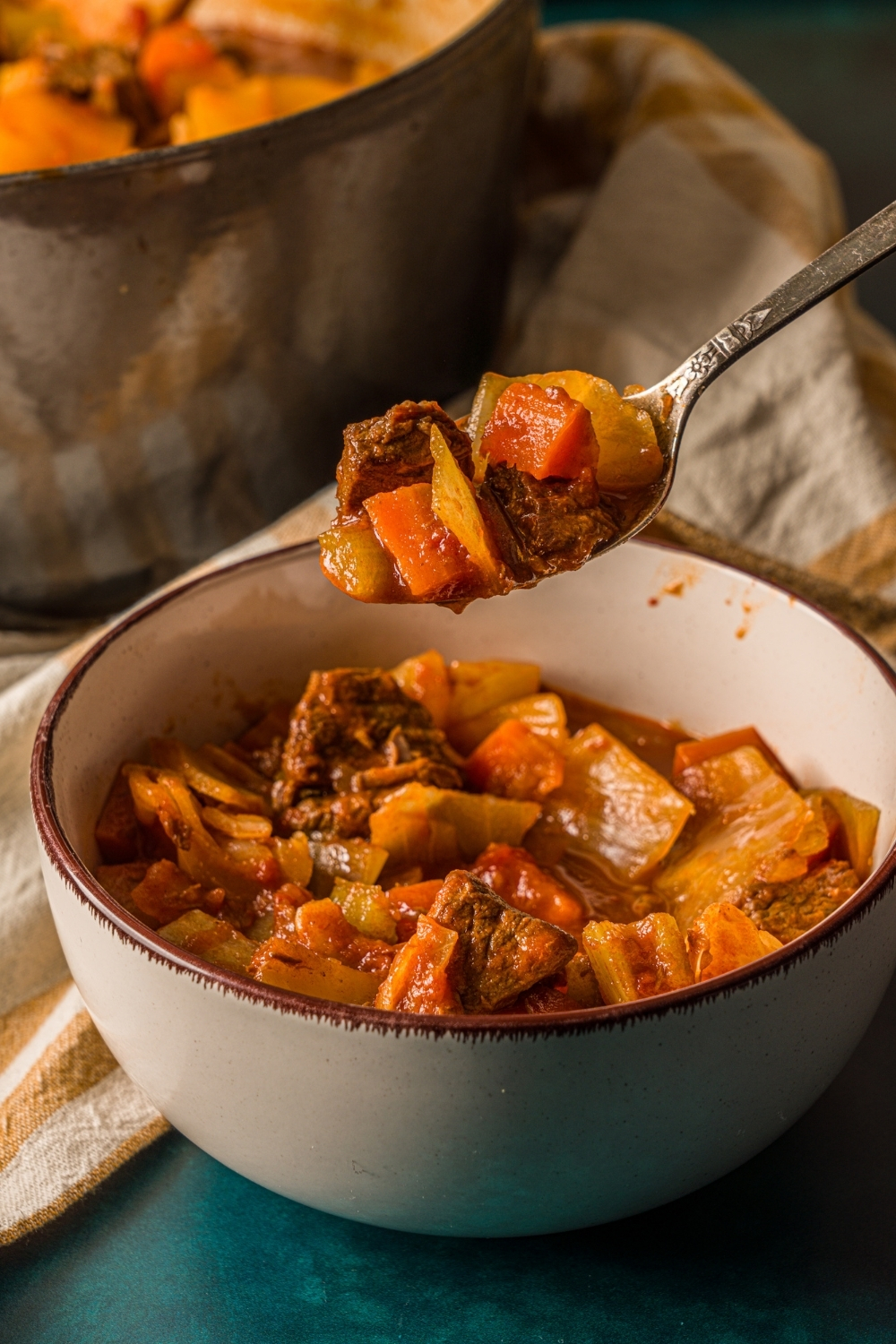 A bowl of beef cabbage stew on a blue counter with a striped cloth napkin. There is a spoon with a bite of stew over the bowl.
