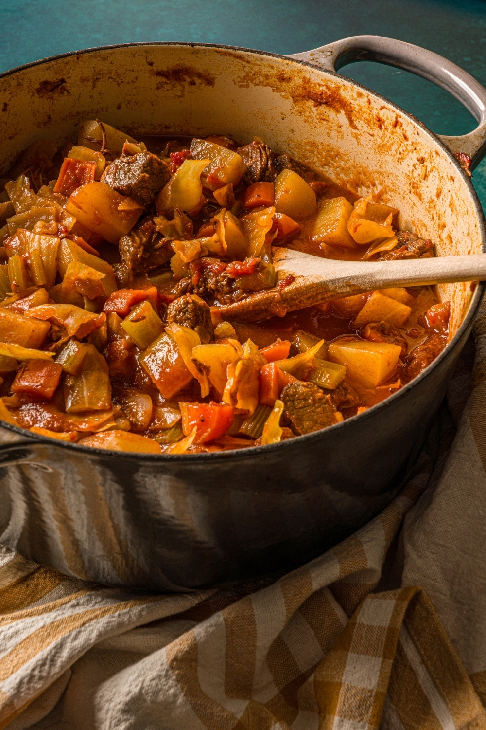 A dutch oven with beef cabbage stew on a blue counter with a striped napkin. A wooden spoon is stirring the stew.