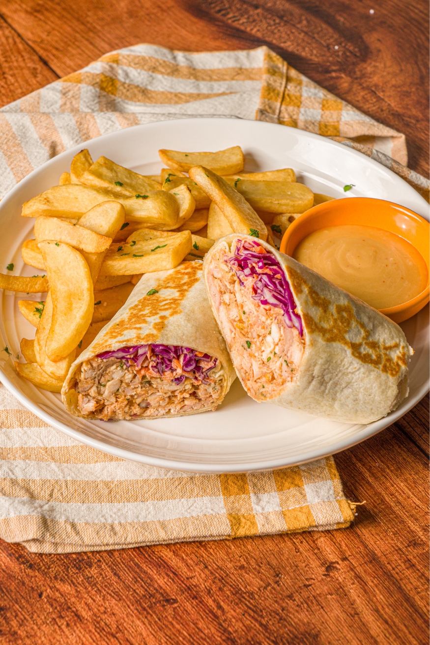 A BBQ chicken wrap sliced in half on a white plate with french fries garnished with fresh parsley. The wrap is served with a small bowl of dipping sauce. The plate is on a wood counter with a yellow striped napkin.