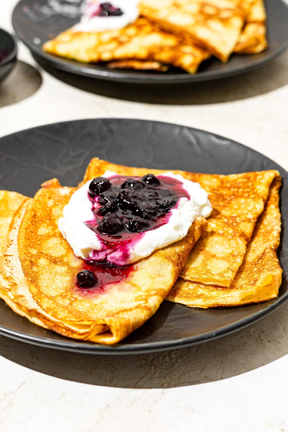 A black palate with Swedish pancakes topped with whipped cream and blueberry jam. The plate is on a white counter with an additional plate of pancakes.