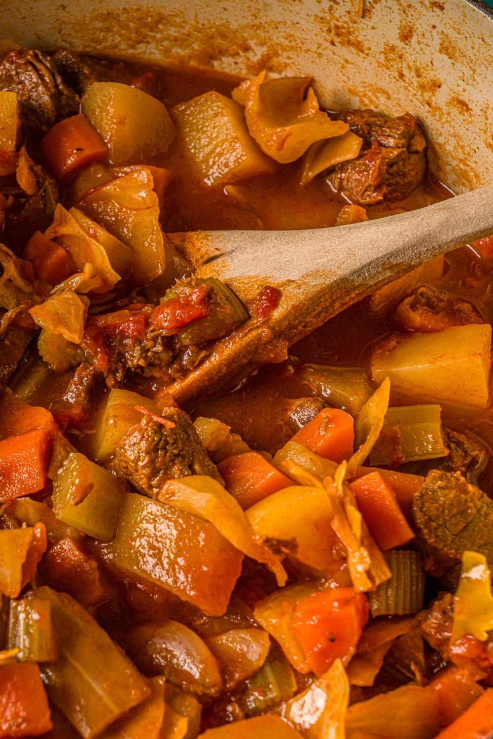 A wooden spoon stirring beef cabbage stew in a dutch oven.