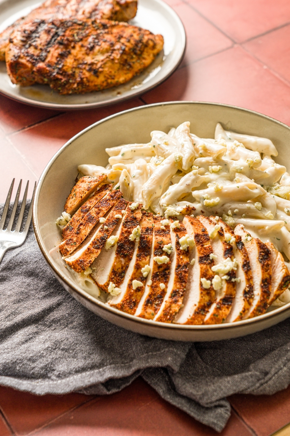 A bowl of blackened chicken alfredo with penne pasta garnished with bleu cheese. The bowl is on a red counter with a gray cloth napkin and fork. There is a plate of chicken on the counter.