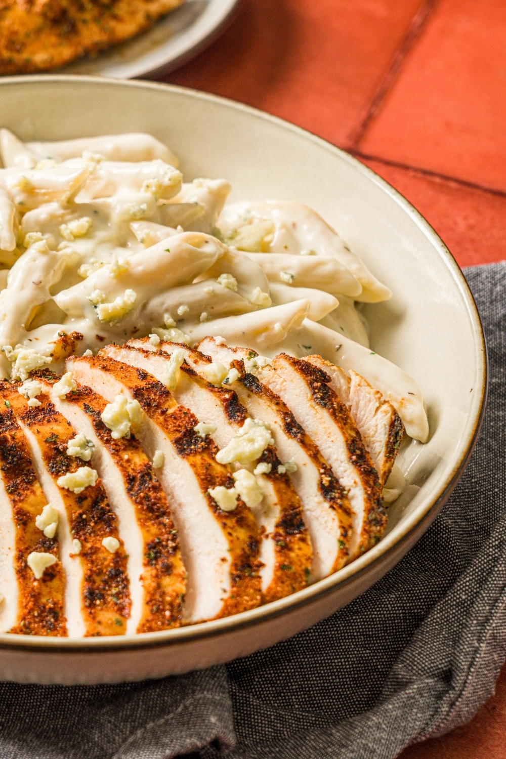 A bowl of blackened chicken alfredo with penne pasta garnished with bleu cheese. The bowl is on a red counter with a gray cloth napkin.