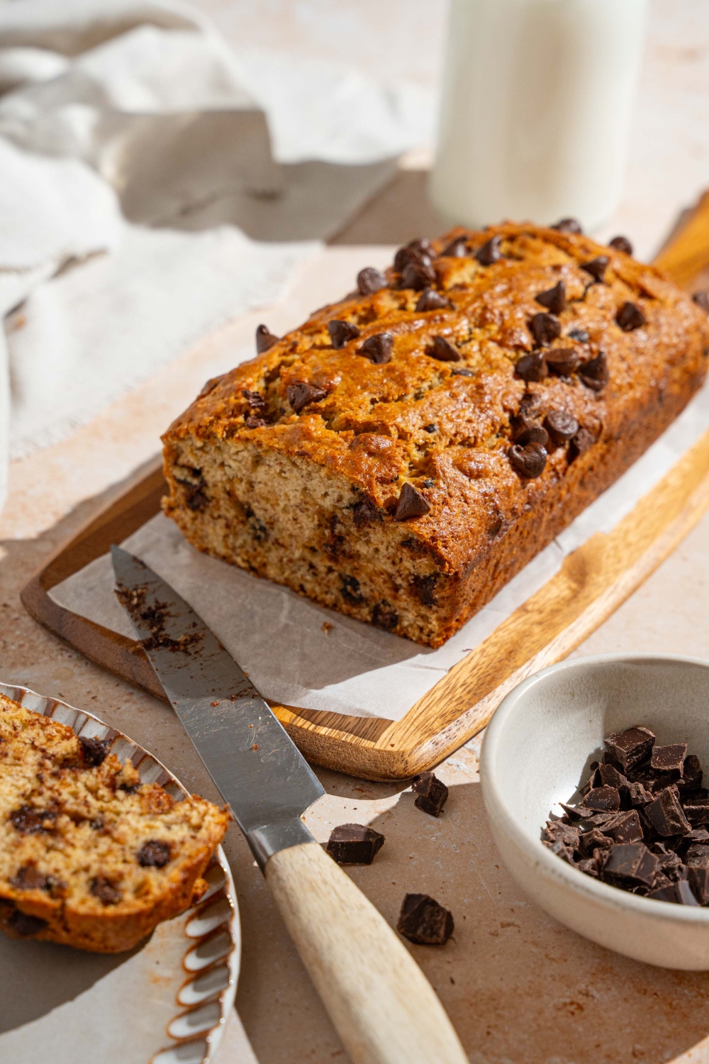 A wooden board lined with parchment paper with brown butter chocolate chip banana bread with a slice removed. The board is on a tan counter with a knife and small bowl of chocolate chips and glass of milk.