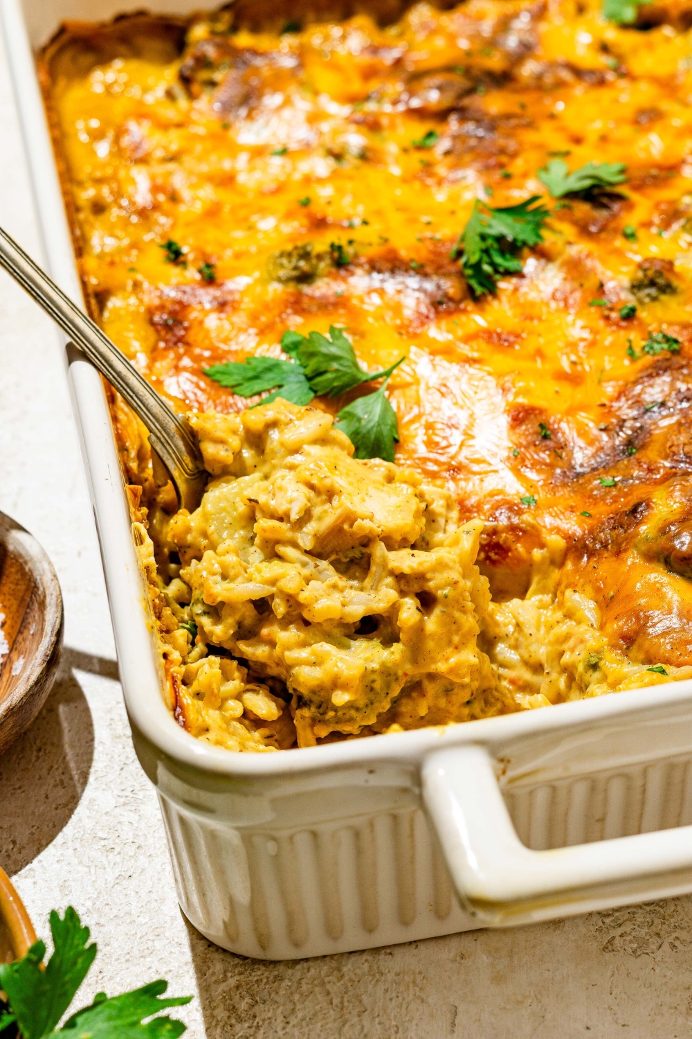 A baking dish with baked chicken broccoli rice casserole garnished with fresh parsley. A spoon is taking a portion of casserole from the dish.