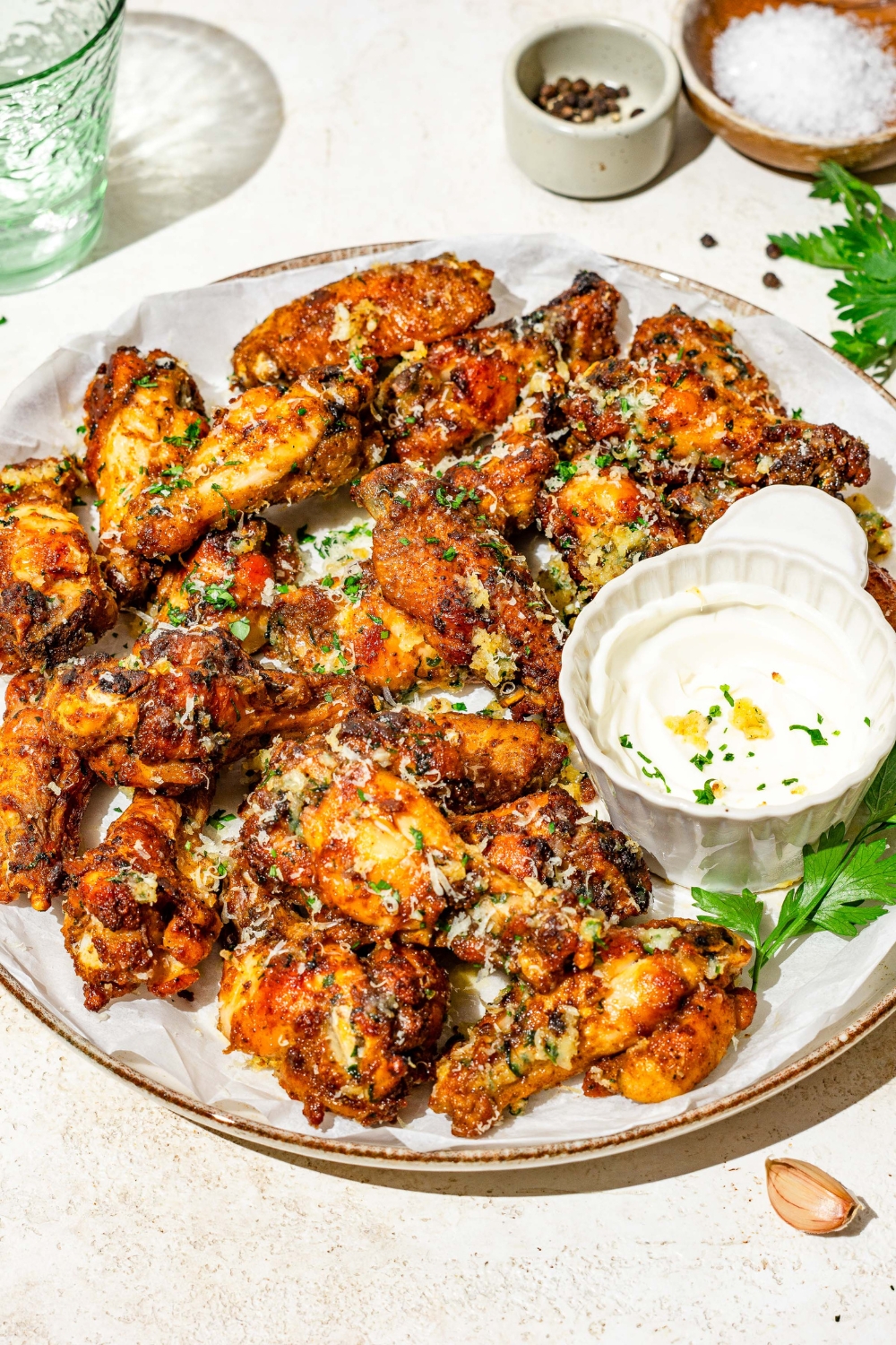 A plate of copycat Wingstop garlic parmesan chicken wings garnished with grated cheese and fresh parsley. There is a bowl of sauce on the plate. The plate is on a tan counter with small bowls of seasonings.