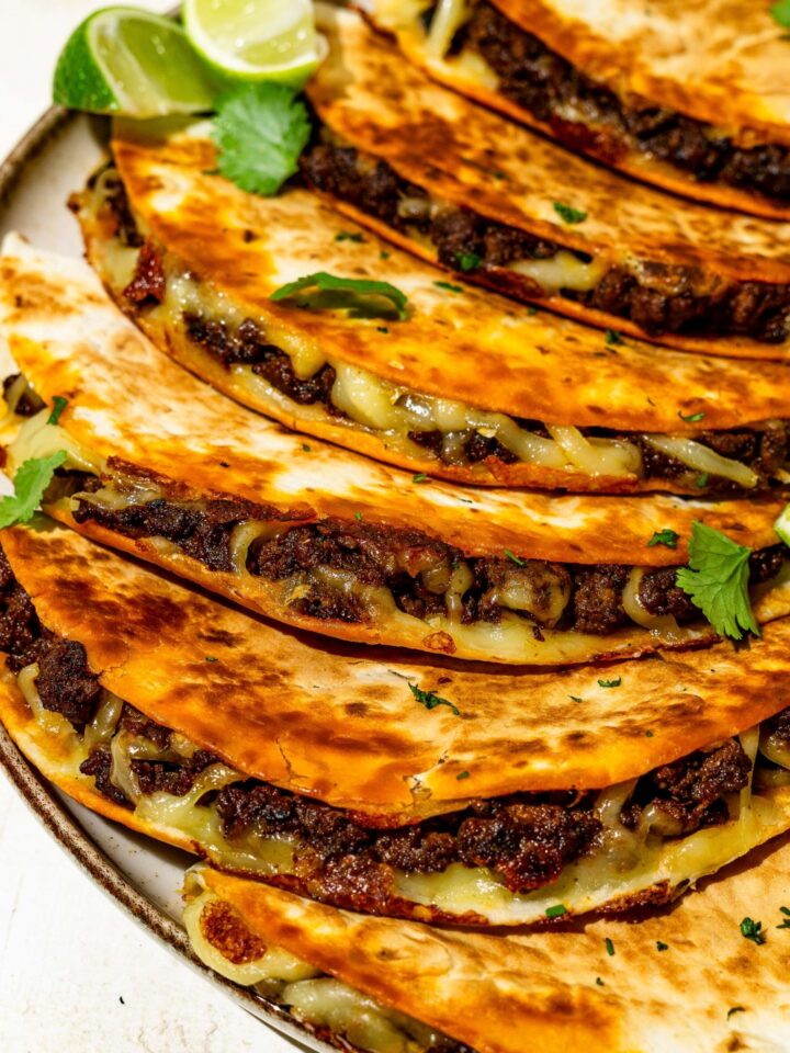 Several crispy beef tacos layered on a plate and garnished with lime and cilantro. The plate is on a tan counter.