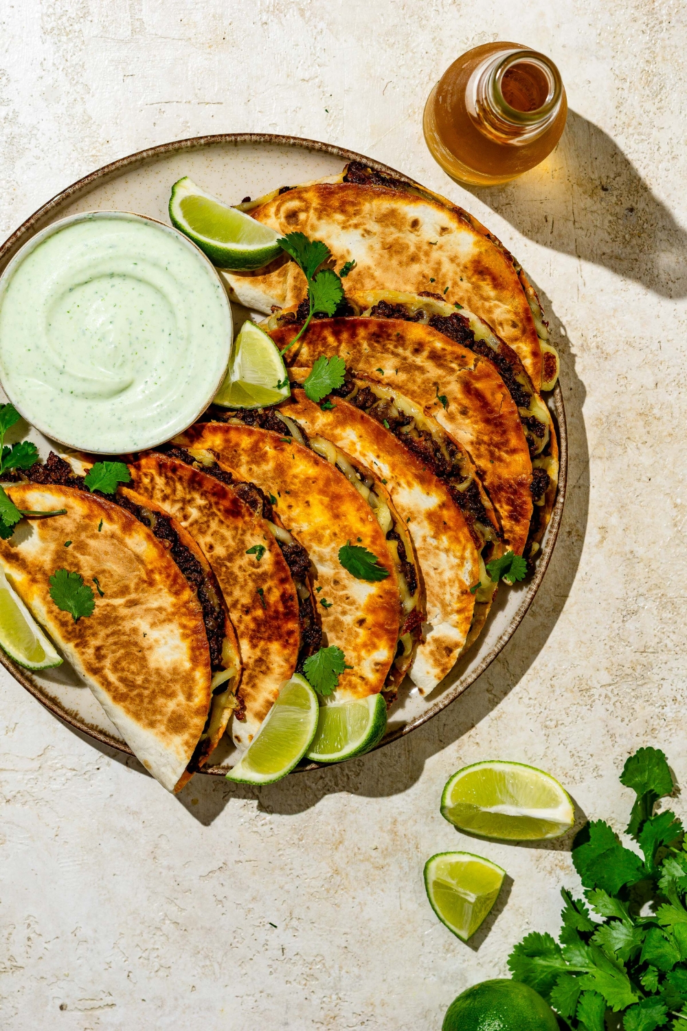 Crispy beef tacos layered on a plate garnished with cilantro and lime wedges and served with a side of sauce. The plate is on a tan counter with garnishes.