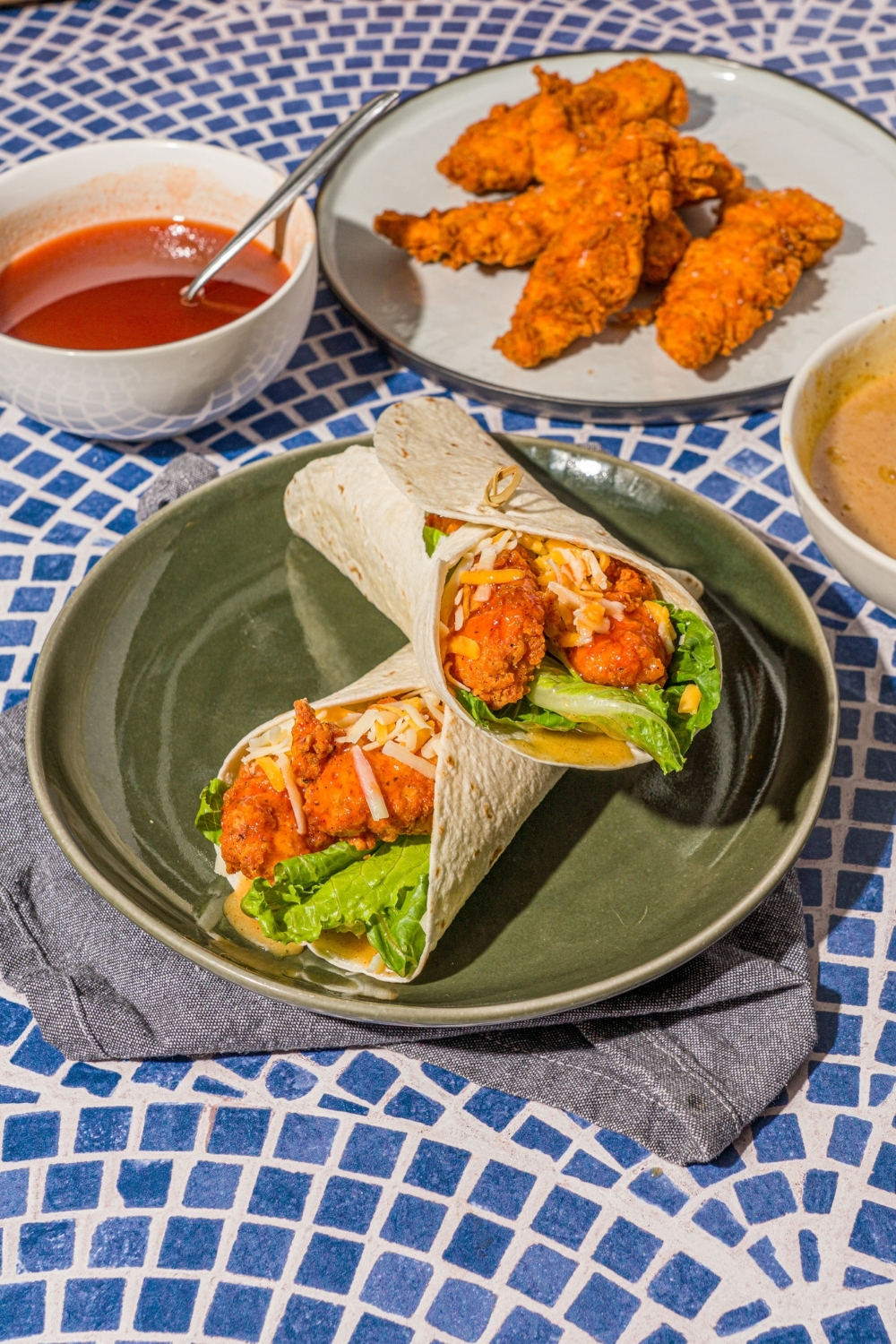 A green plate with two crispy chicken wraps stacked on top of the other. The plate is on a gray cloth napkin on a blue tiled counter with a plate of chicken tenders and a bowl of hot honey sauce.
