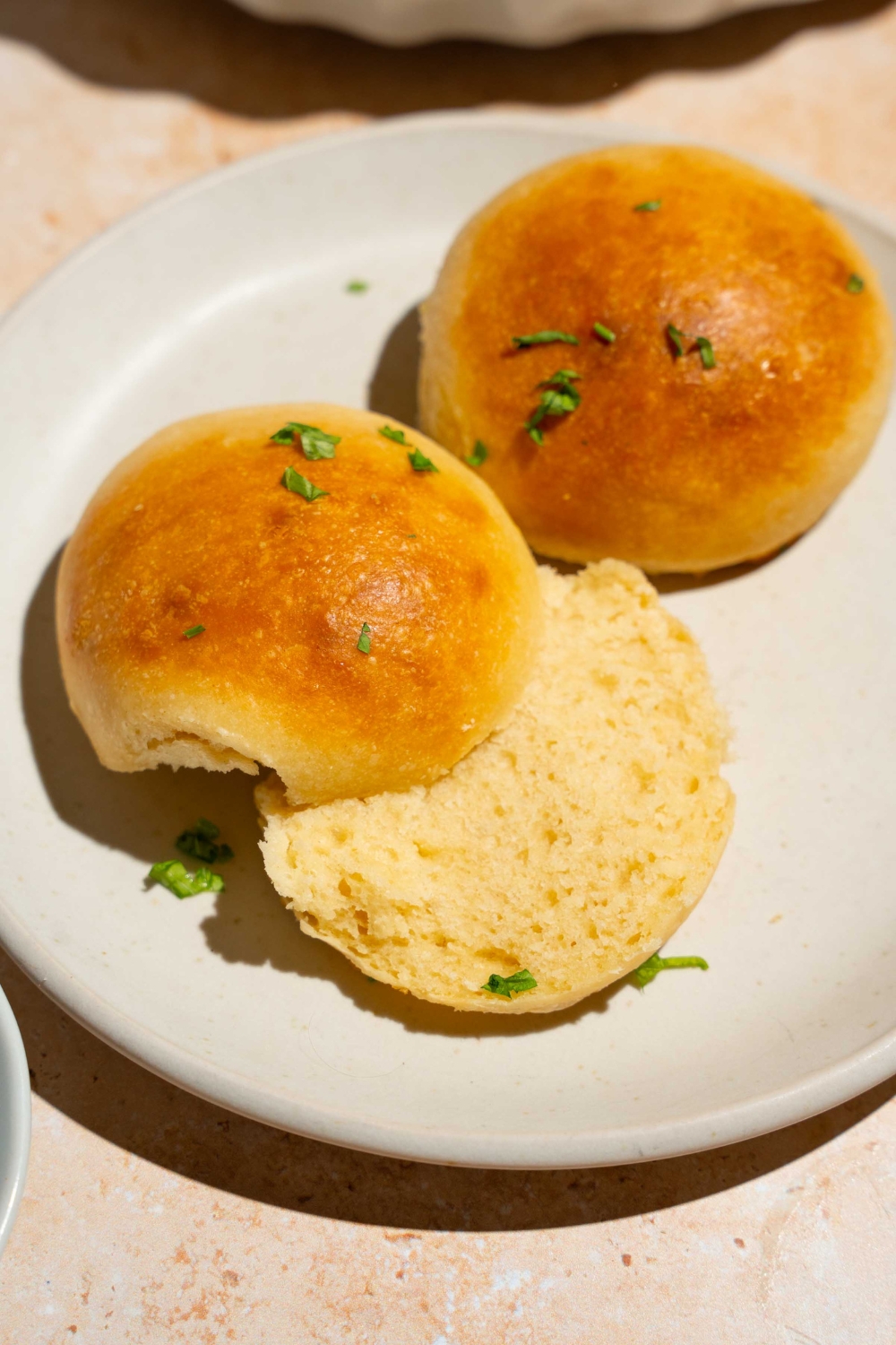 A white plate with two dinner rolls brushed with garlic herb butter. One of the rolls is sliced in a half. The plate is on a tan counter.