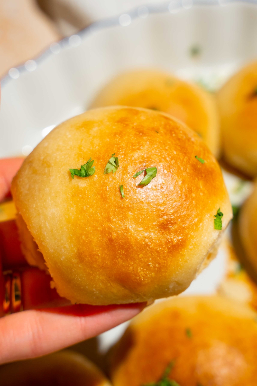 A close up of a hand holding a dinner roll brushed with herb butter. There is a baking dish of rolls blurred in the background.