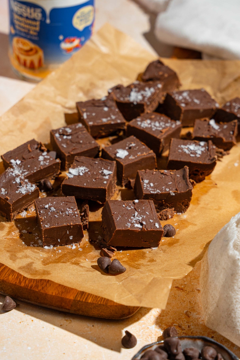 Sliced fudged pieces topped with flaky sea salt on a wooden board lined with parchment paper. The board is on a tan counter with a can of condensed milk and small bowl of chocolate chips.
