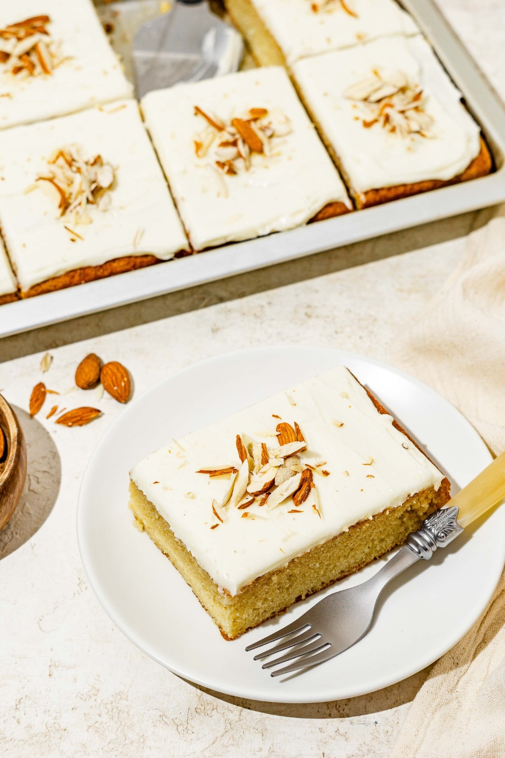 A white plate with a slice of white Texas sheet cat topped with sliced almonds. There is a fork on the plate. The plate is on a white counter with a baking pan with cake.