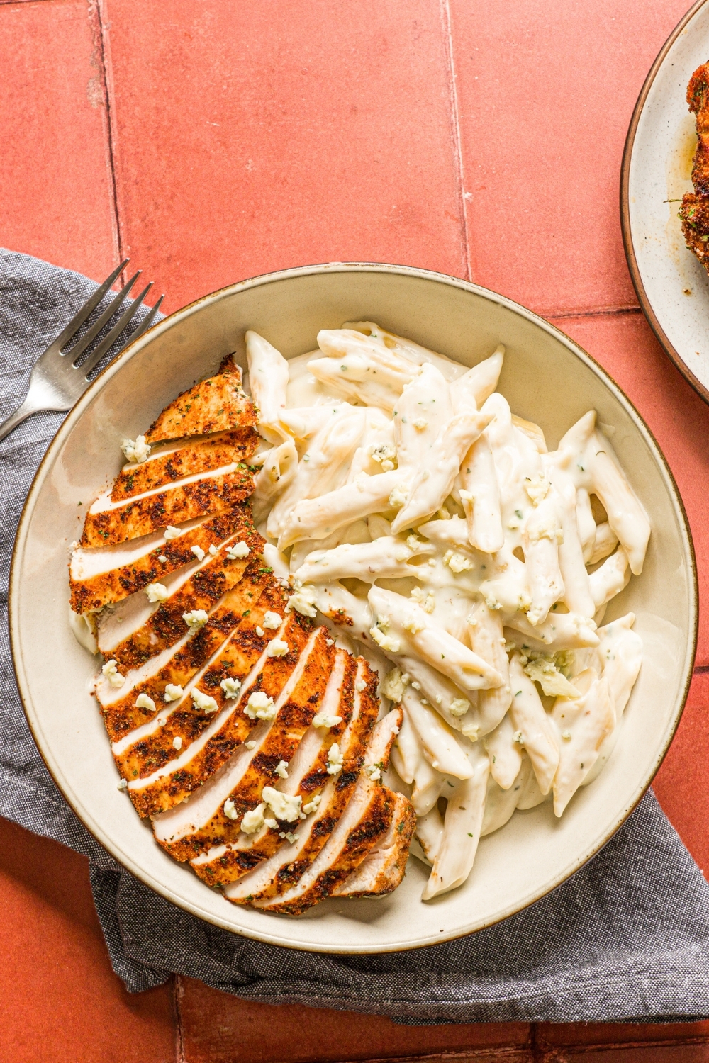 A bowl of blackened chicken alfredo with penne pasta garnished with bleu cheese. The bowl is on a red counter with a gray cloth napkin and fork.