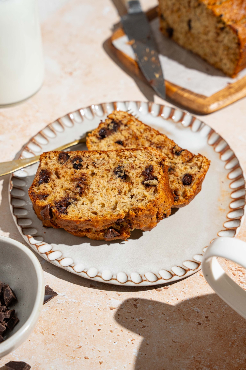 Two slices of brown butter chocolate chip banana bread on a white plate. The plate is on a tan counter with a board of banana bread with a knife.