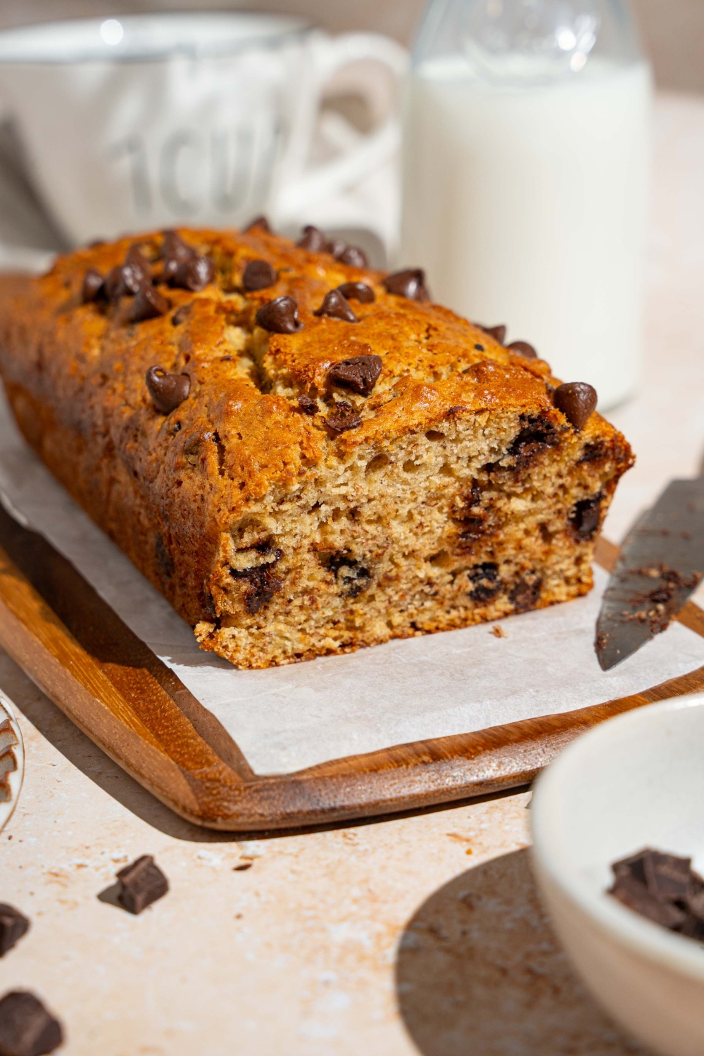 A wooden board lined with parchment paper with brown butter chocolate chip banana bread with a slice removed. The board is on a tan counter with a knife and small bowl of chocolate chips and glass of milk.