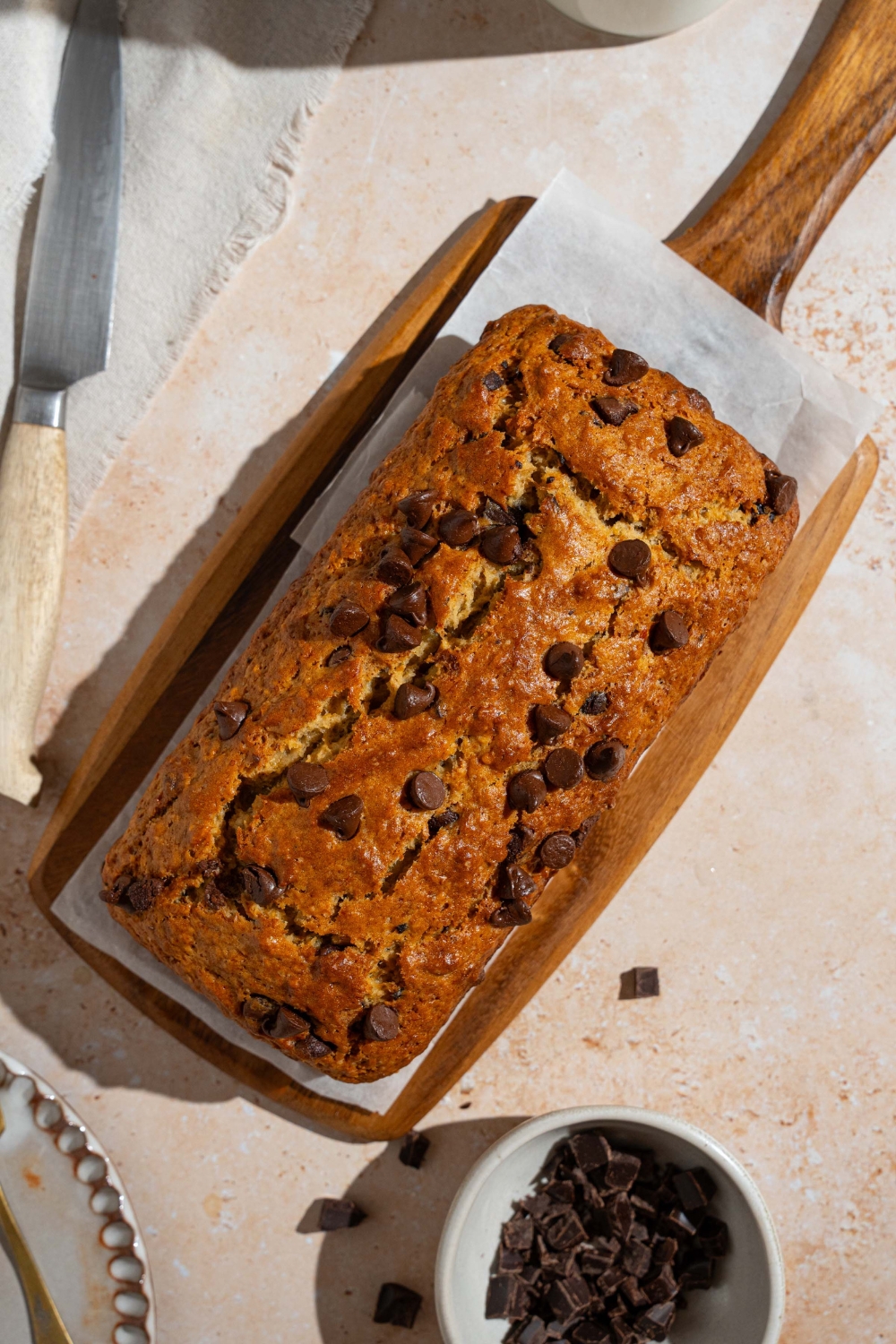 A wooden board lined with parchment paper with baked brown butter chocolate chip banana bread. The board is on a tan counter with a small bowl of chocolate chips.