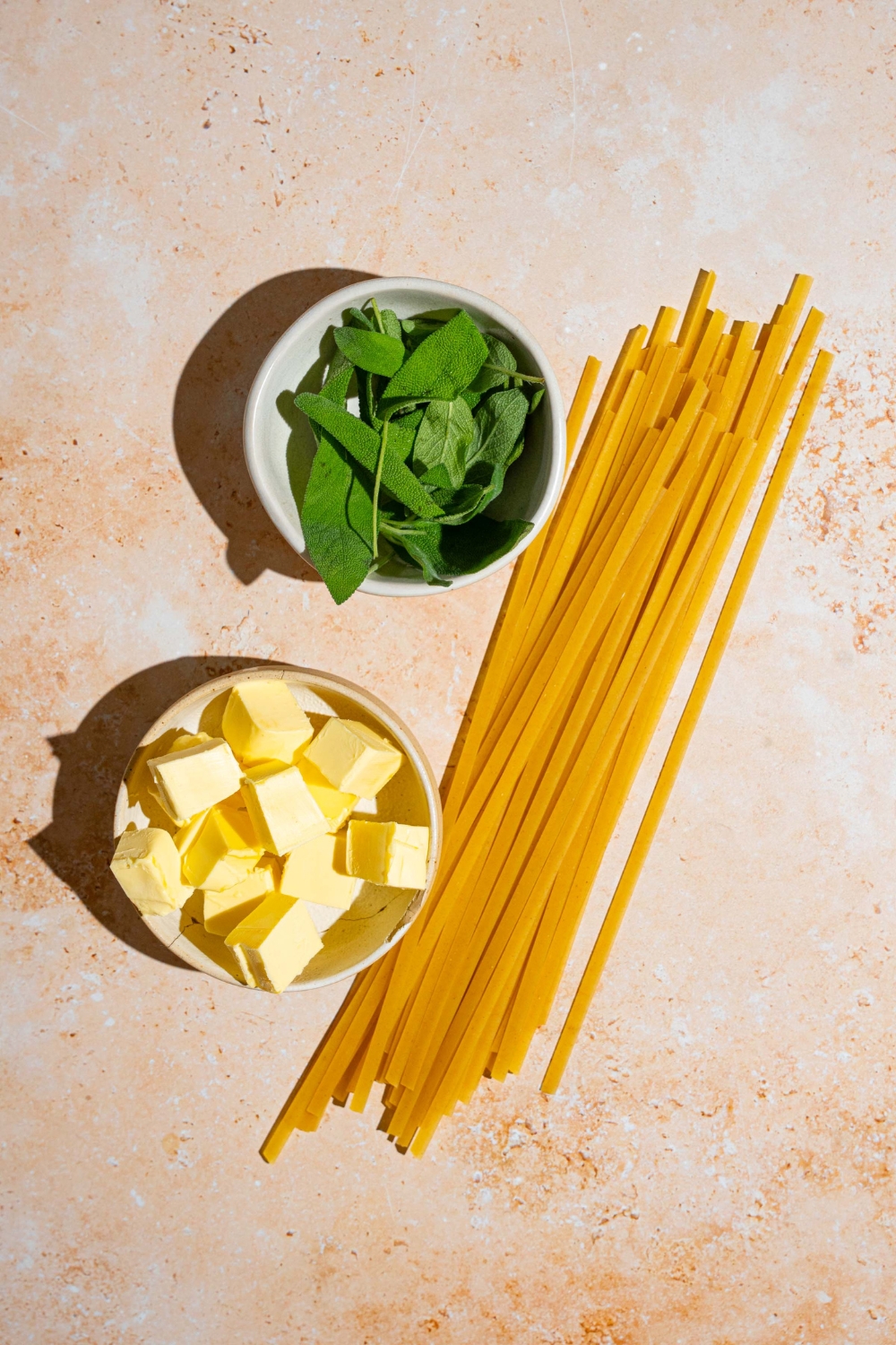 Ingredients to make brown butter sauce including uncooked linguine pasta, a bowl of sage, and a bowl of cubed butter.
