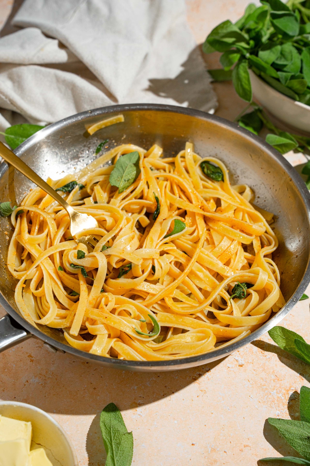 A skillet with linguine pasta tossed in brown butter sauce and sage. There is a fork in the skillet. The skillet is on a tan counter with a bowl of sage and white cloth napkin.