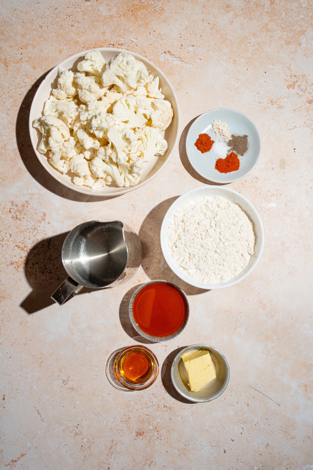 An overhead shot of several bowls in various sizes containing ingredients to make Buffalo cauliflower wings including cauliflower florets, hot sauce, butter, milk, and seasonings.