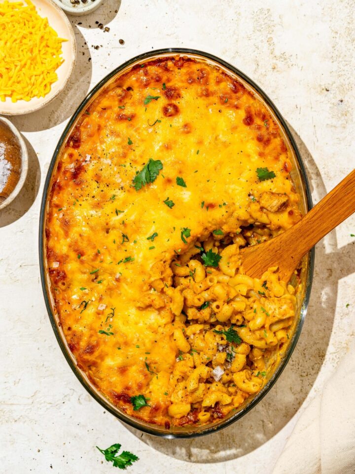 An oval glass baking dish with baked Buffalo mac and cheese garnished with fresh parsley. There is a wooden spoon scooping mac and cheese from the dish. The baking dish is on a tan counter with small plates of garnishes.