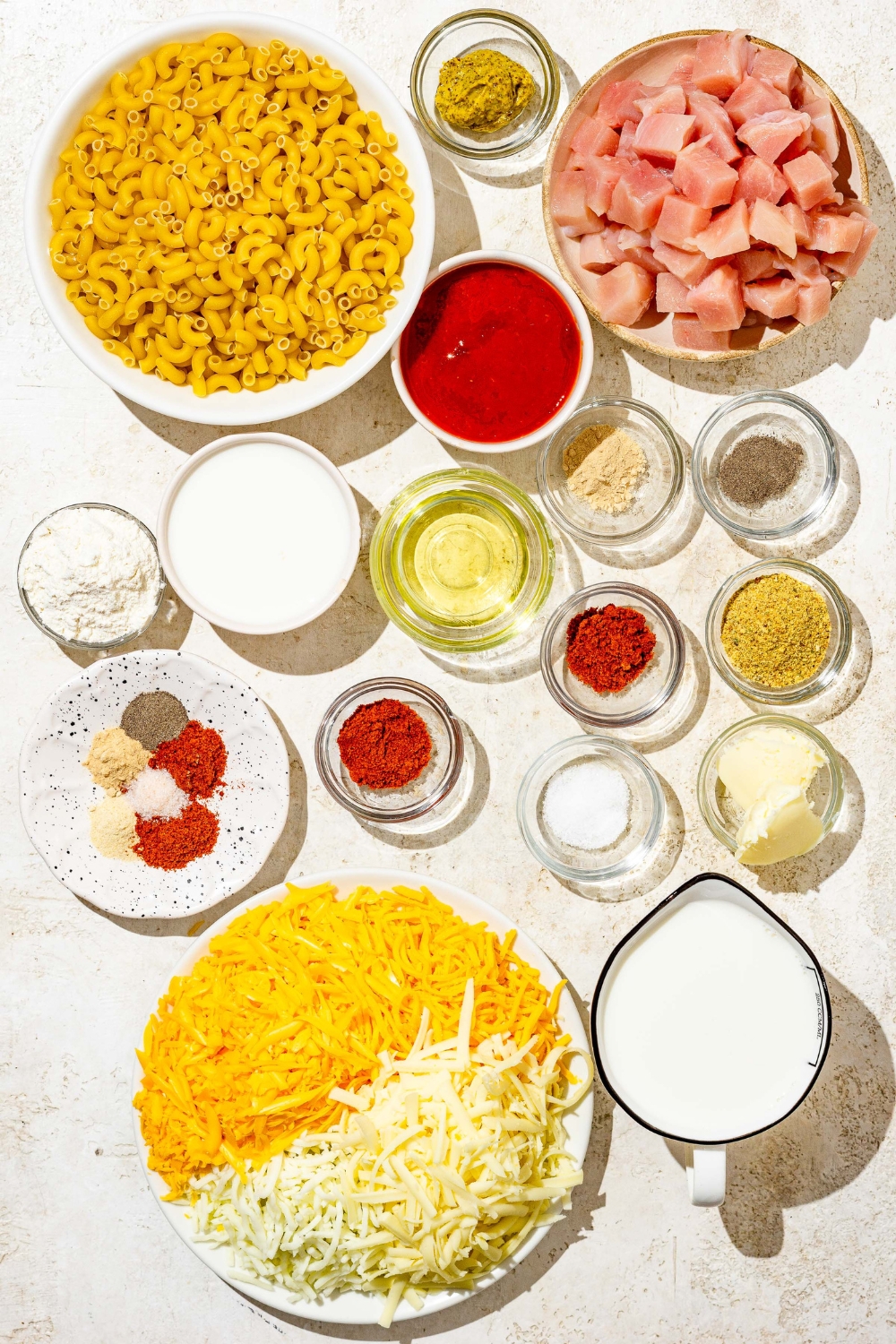 An overhead shot of several bowls in various sizes containing ingredients to make Buffalo mac and cheese including cubed chicken, elbow pasta, shredded cheeses, milk, cream, flour, butter, dijon mustard, hot sauce, and seasonings.