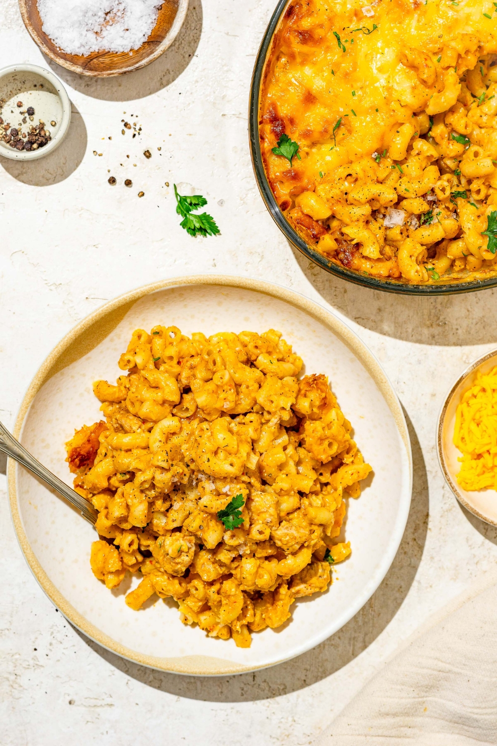 A plate of Buffalo mac and cheese garnished with fresh parsley and served with a fork. The plate is on a tan counter with a baking dish fo mac and cheese and small plates of garnishes.