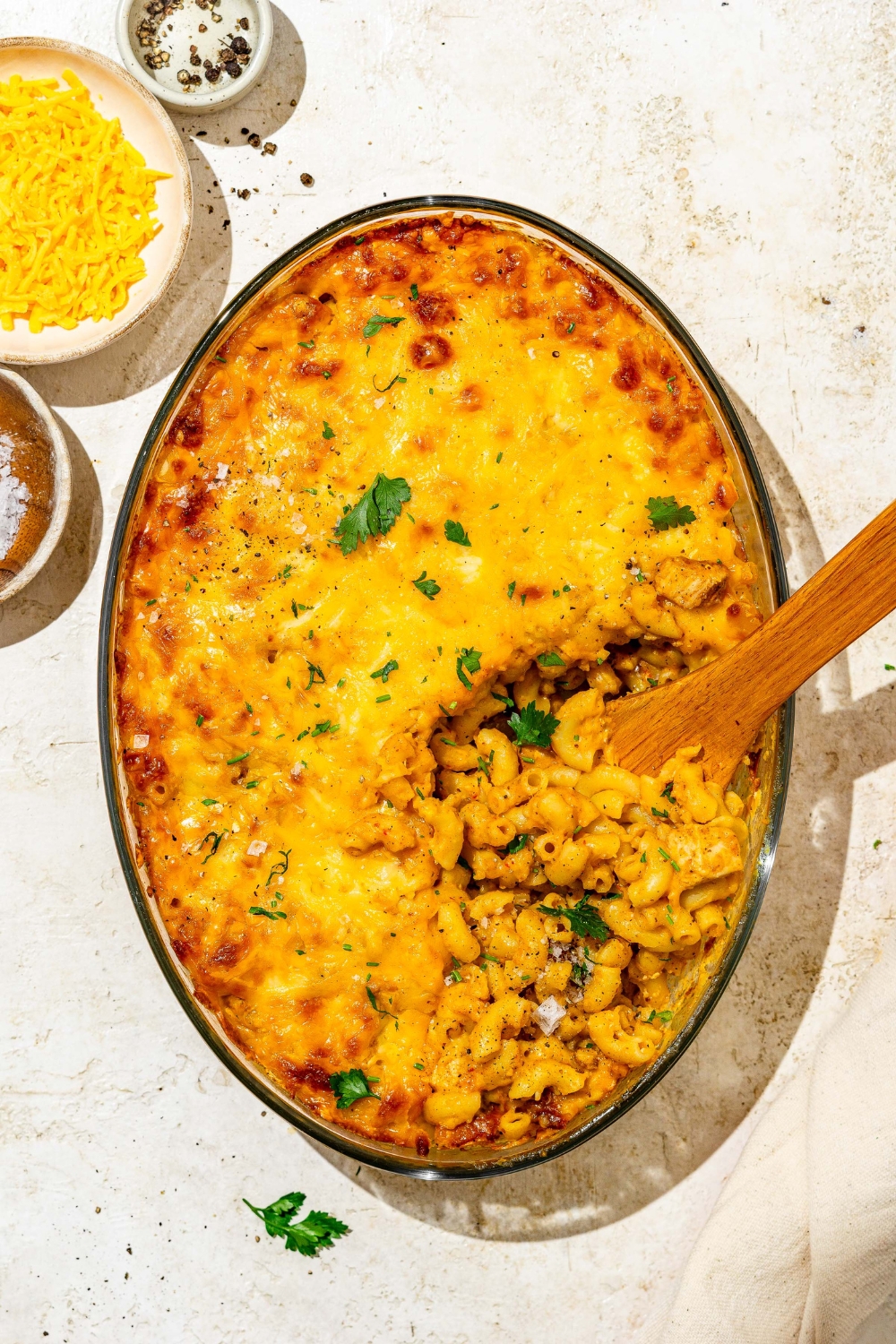 An oval glass baking dish with baked Buffalo mac and cheese garnished with fresh parsley. There is a wooden spoon scooping mac and cheese from the dish. The baking dish is on a tan counter with small plates of garnishes.
