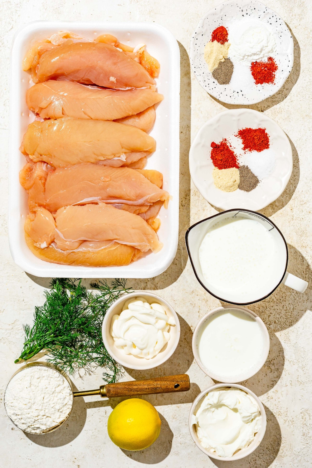 An overhead shot of several bowls containing ingredients to make buttermilk fried chicken tenders including chicken tenders, buttermilk, flour, mayonnaise, sour cream, fresh dill, lemon, and seasonings.