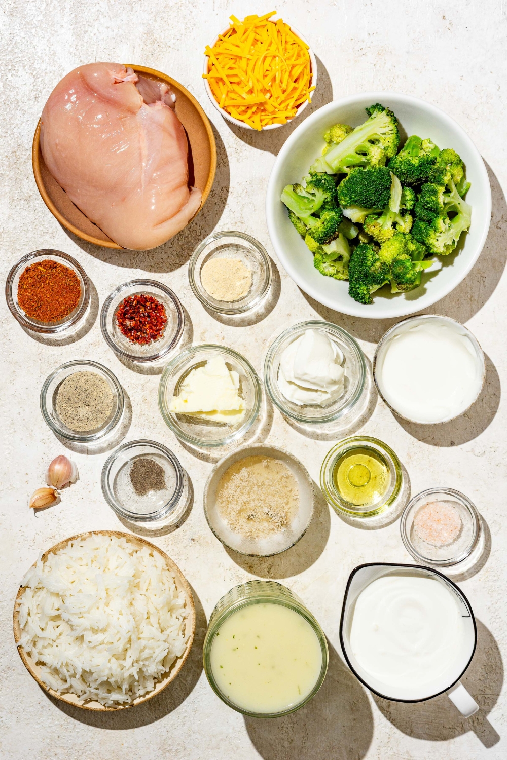 An overhead shot of several bowls in various sizes containing ingredients to make chicken broccoli rice casserole including chicken breasts, rice, broccoli, shredded cheese, oil, cream, cream of chicken, cream cheese, garlic, and seasonings.