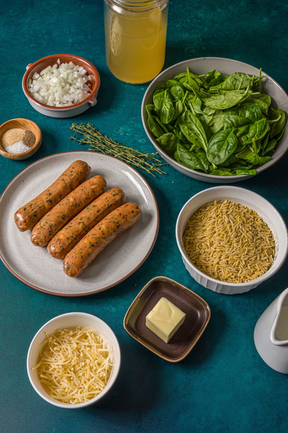 Several bowls and plates in various sizes containing ingredients to make chicken sausage orzo including orzo pasta, chicken sausage links, spinach, grated cheese, butter, diced onion, cream, chicken broth, and seasonings.