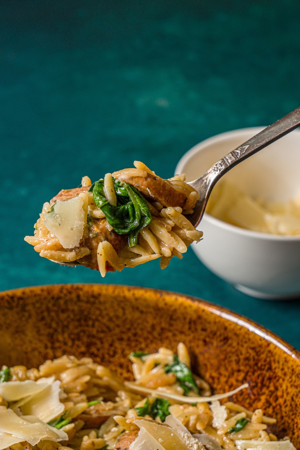 A spoon with a bite of chicken sausage orzo over a bowl of orzo. The bowl is on a blue counter with a small bowl of shaved parmesan.