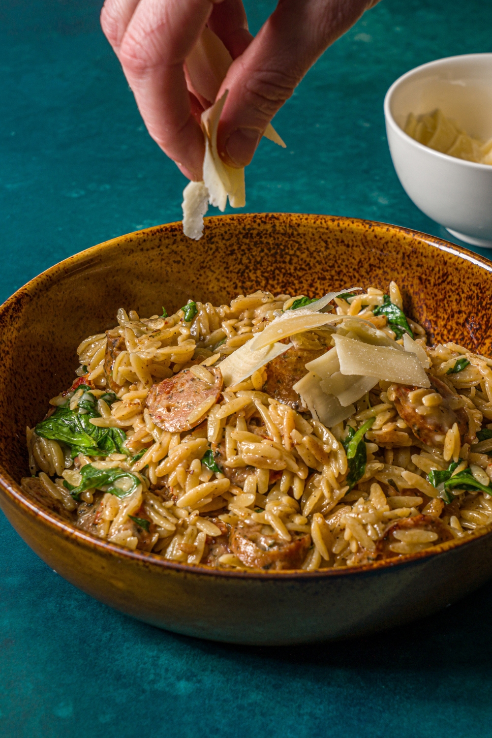 A bowl of chicken sausage orzo pasta with a hand sprinkling shaved parmesan over the bowl. The bowl is on a blue counter with a small bowl of shaved parmesan.