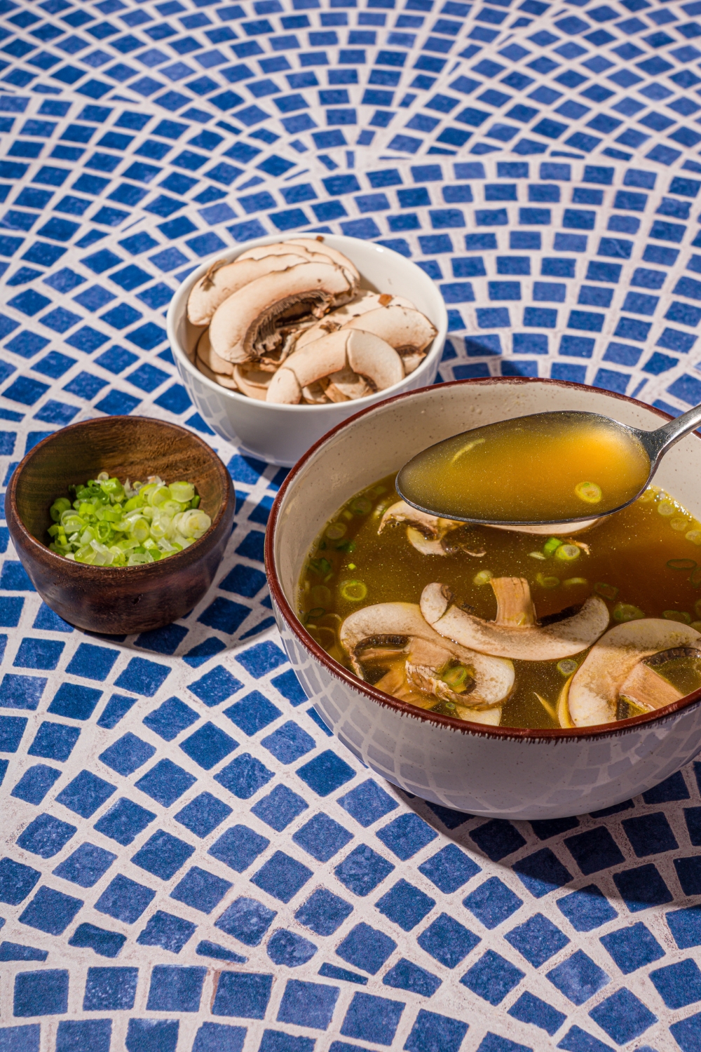A ceramic bowl with clear soup with sliced mushrooms and green onions on a blue tiled counter with a small bowl of sliced mushrooms. There is a spoon scooping a bite of soup.