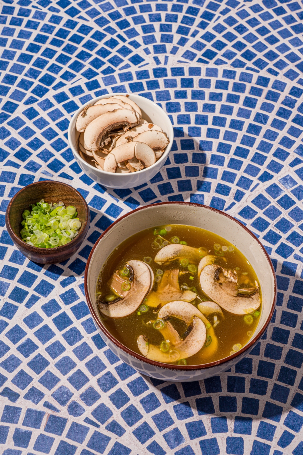 A ceramic bowl with clear soup with sliced mushrooms and green onions on a blue tiled counter with a small bowl of sliced mushrooms.