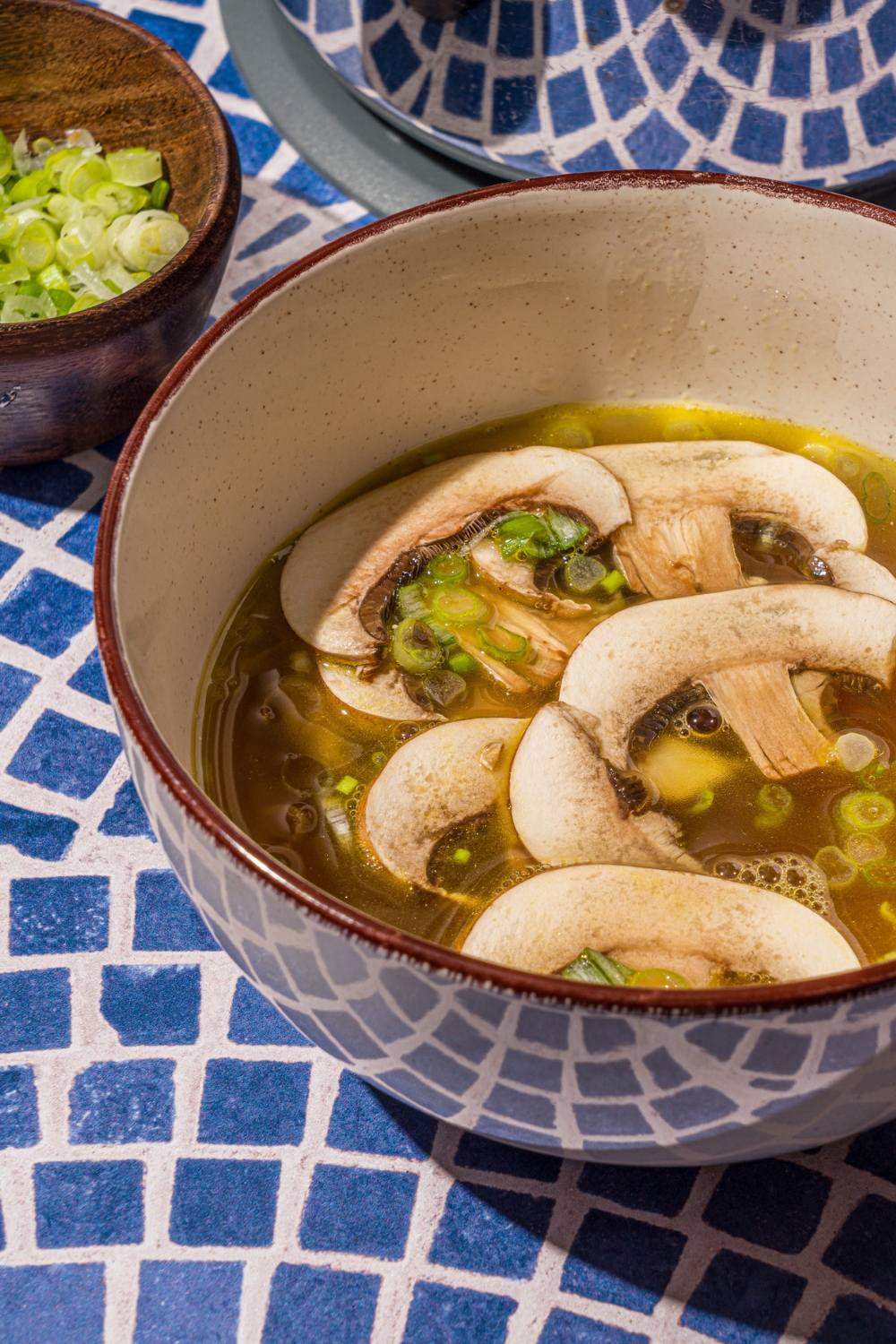 A ceramic bowl with clear soup with sliced mushrooms and green onions on a blue tiled counter with a small bowl of sliced mushrooms.
