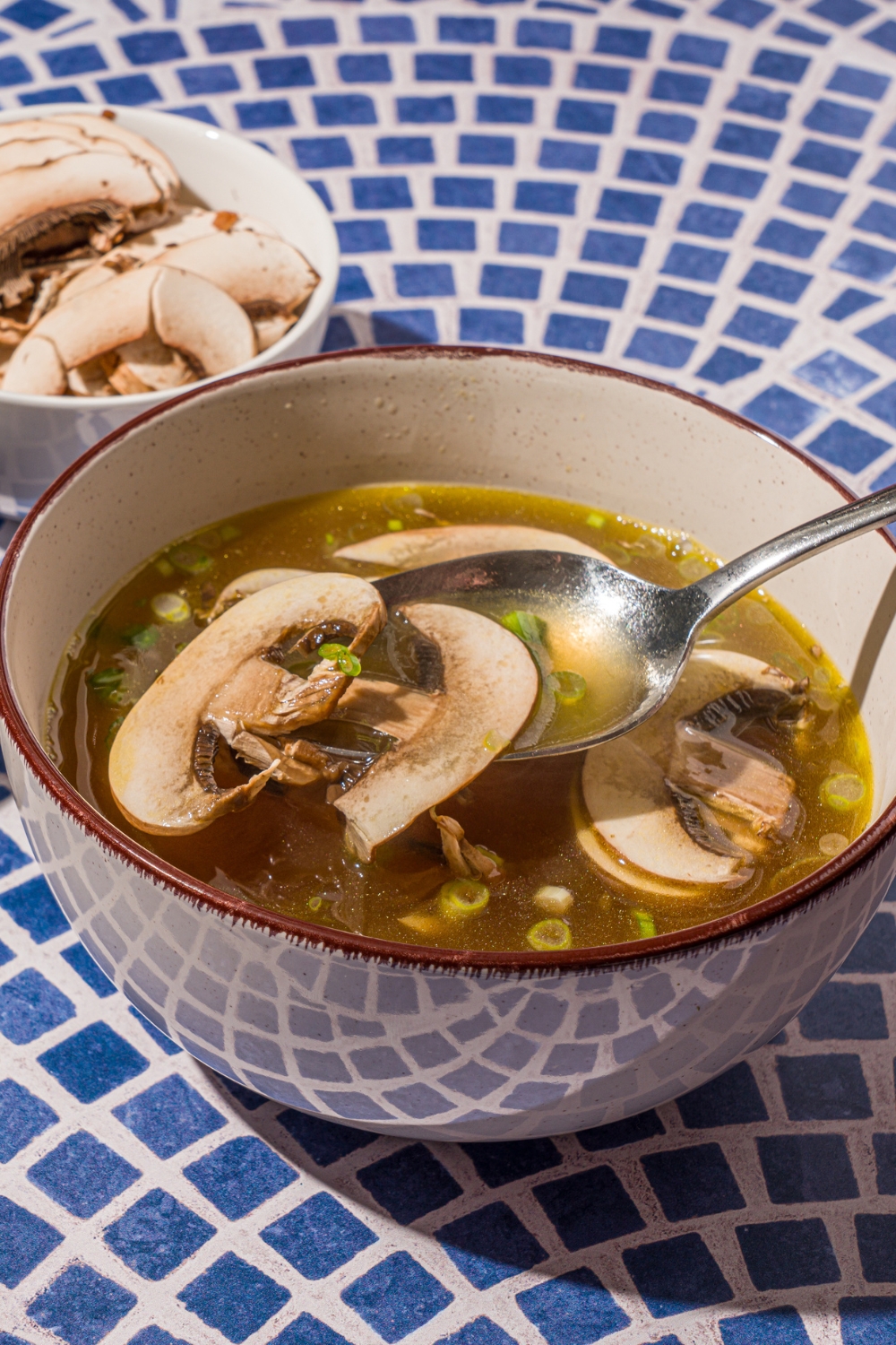 A ceramic bowl with clear soup with sliced mushrooms and green onions on a blue tiled counter with a small bowl of sliced mushrooms. There is a spoon scooping a bite of soup.