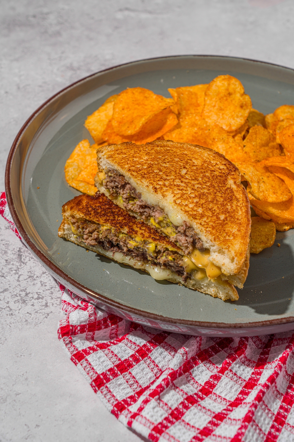 A green plate with a sliced copycat Applebee's grilled cheeseburger stacked on top of each other. There are chips on the plate. The plate is on a stone counter with a red checkered napkin.