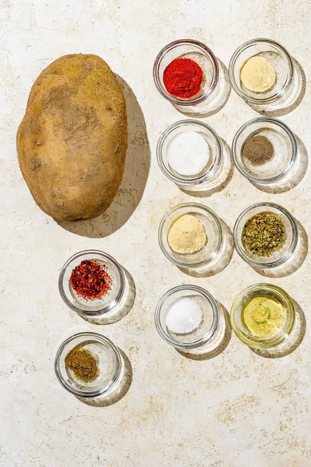 An overhead shot of a potato and several bowls containing ingredients to make copycat Five Guys cajun fries including sugar, smoked paprika, salt, pepper, garlic powder, onion powder, oregano, crushed red pepper flakes, and thyme.