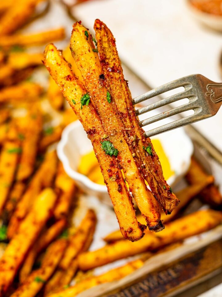 A close up of a fork with three copycat Five Guys cajun fries garnished with fresh parsley. There is a baking sheet of fries on a white counter blurred in the background.