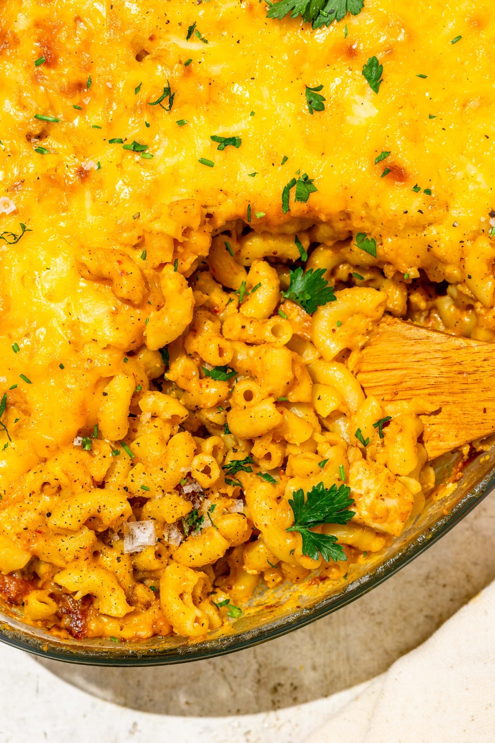 An oval glass baking dish with baked Buffalo mac and cheese garnished with fresh parsley. There is a wooden spoon scooping mac and cheese from the dish.